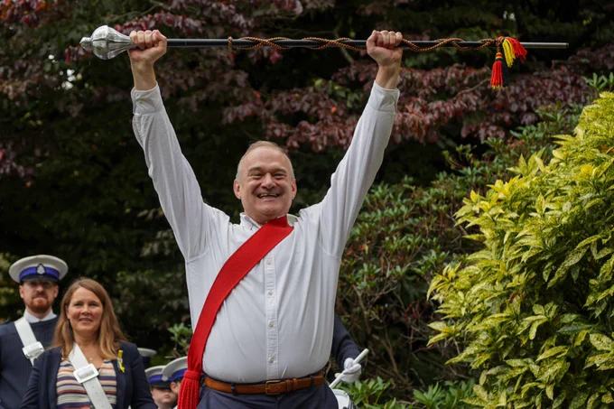 Ed Davey smiles and raises a band leader’s baton above his head