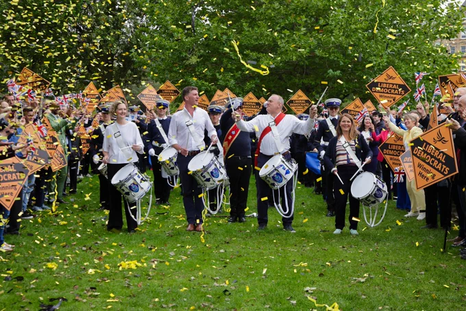 Ed Davey, Edward Morello, Vikki Slade and Anna Sabine play drums at a Liberal Democrat rally with confetti falling around supporters holding signs.