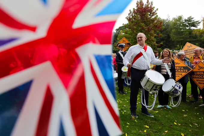 Ed Davey smiling and holding some drums at a Lib Dem rally, with a large Union Jack flag in the foreground.