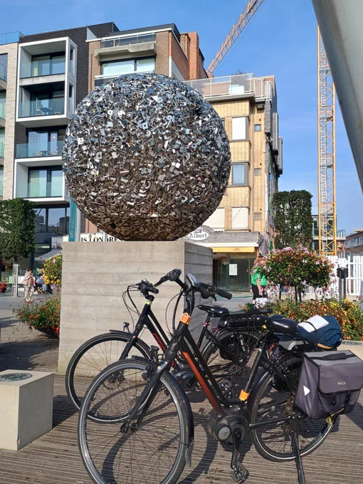 Picture of a large metal  sculpture appearing to be a ' ball of bits ' , from the centre of Koksijde
.
In front are two dutch style electric bikes, behind is an apartment building.