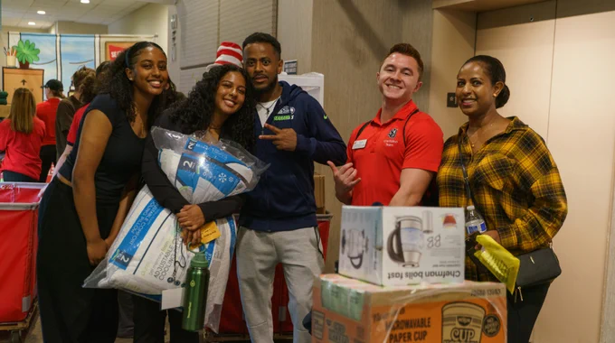 Group of five people smiling in a dorm lobby, holding various household items like pillows and kitchen appliances.
