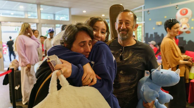 Two people hugging, with another holding a stuffed toy and smiling beside them in a lively indoor setting.