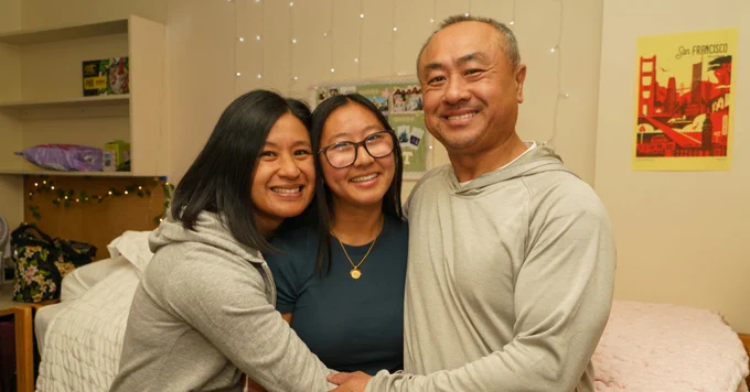 Three individuals posing happily in a dorm room with a string of lights and a "San Francisco" poster in the background.
