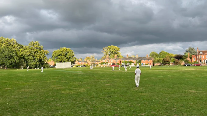 View from the boundary shows brooding cloud above cricketers watching on as batter tries to hit ball and the umpire looks on. Red brick houses in sunshien can be seen in the background