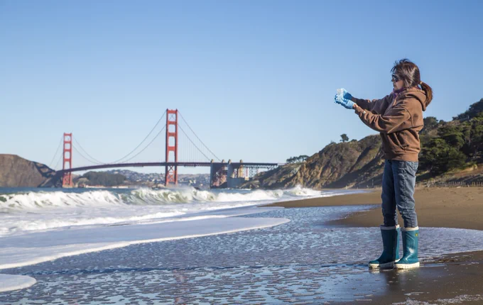 Person wearing blue rain boots and a brown hoodie stands on a sandy beach near the waves with the Golden Gate Bridge in the background.