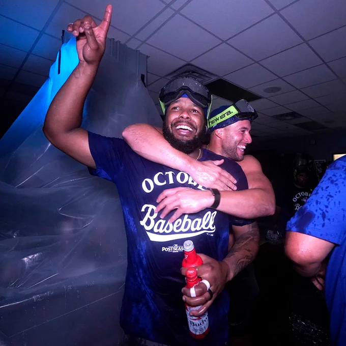 George Springer hugs Vladimir Guerrero Jr. during a clubhouse celebration after the Blue Jays clinch a postseason spot. They are wearing soaked blue October Baseball t-shirts.