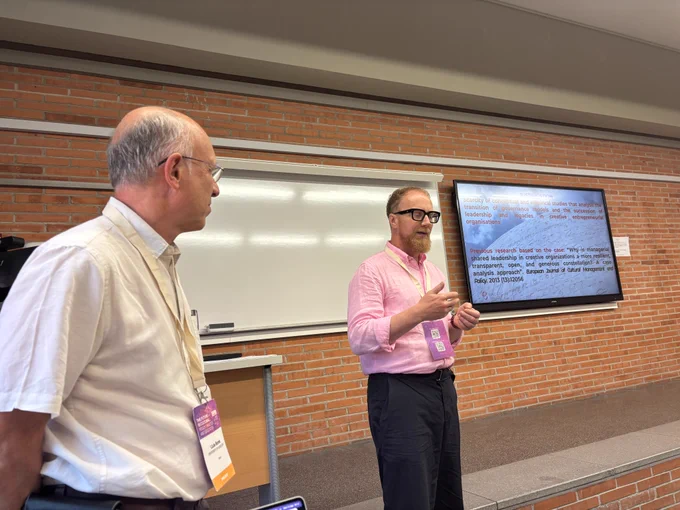 Two men present to a room with a powerpoint showing on a screen behind them. Anders Rykkja wears a pink shirt, Lluis Bonet of Univ of Barcelona is in a white shirt beside him.