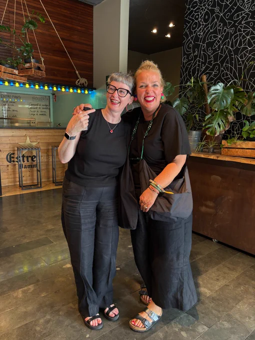 Two women stand laughing. Both are dressed in black with black sandals.