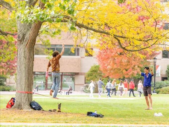 Person balancing on a slackline on the Ag Quat at Cornell with vibrant autumn trees, while another person observes and enjoys the scene. There are lush green and yellow leaves, and students can be seen in the background walking across the Ag Quad at Cornell University.