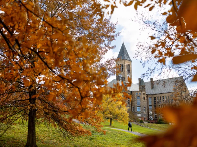 Autumn foliage frames a view of McGraw Tower on the Cornell University campus, with a person walking along the Cornell University Libe Slope.