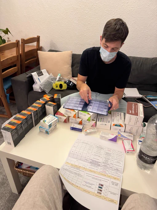 Luke is wearing a face mask, whilst sorting out all of his medication for post transplant. The table is full of boxes all lined up and pieces of paper spread across the table. 