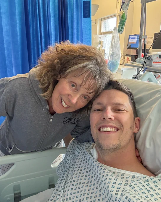 Luke, a male with short hair, is posing for a selfie with his mum, Gill, a woman with shoulder length curly hair, who is leaning in close from the side.

Luke is lying down in his a hospital bed with a white gown on and Gill is in a grey jumper. 