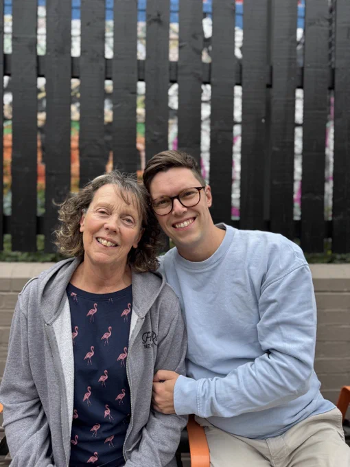 Gill and Luke smiling at the camera. They are sitting outsit. Luke is holding is mums arm in a loving way. Gill is wearing a blue top with flamingos, and a grey hoodie. Luke is wearing a glasses, a blue jumper and light trousers.  