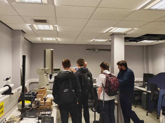 Four individuals with backpacks view a large piece of equipment in a laboratory setting.