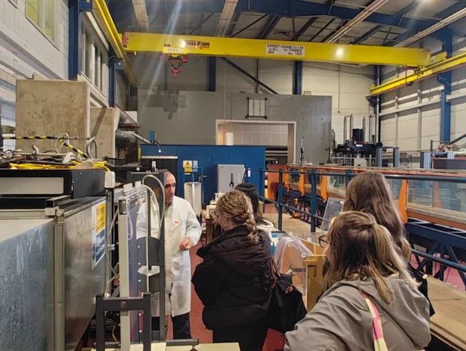 A group of visitors listening to a guide in a lab coat during a tour of an industrial facility with heavy machinery and a yellow overhead crane.