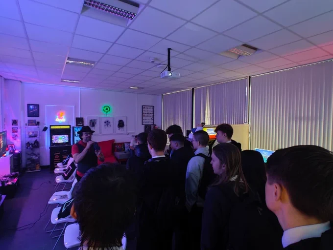 A group of people gathered in a dimly lit room featuring colorful neon lights and arcade games, focusing attention towards a person in the center who appears to be instructing or presenting.