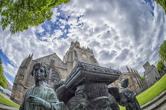 The historic buildings and statues of King's College campus with a cloudy blue sky and green lawn and trees