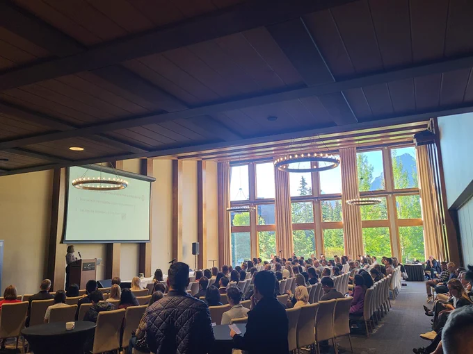 A conference room with floor-to-ceiling windows, people sitting in chairs watching an academic talk on a large screen.