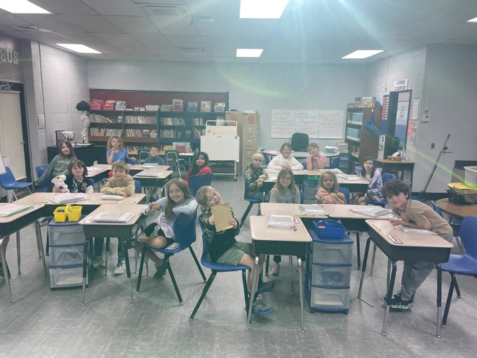 15 students sit at their desks in mrs. jackson's 3rd grade room. they are smiling at the camera. one girl in the front is holding a golden Lego brick and another girl is holding a golden book