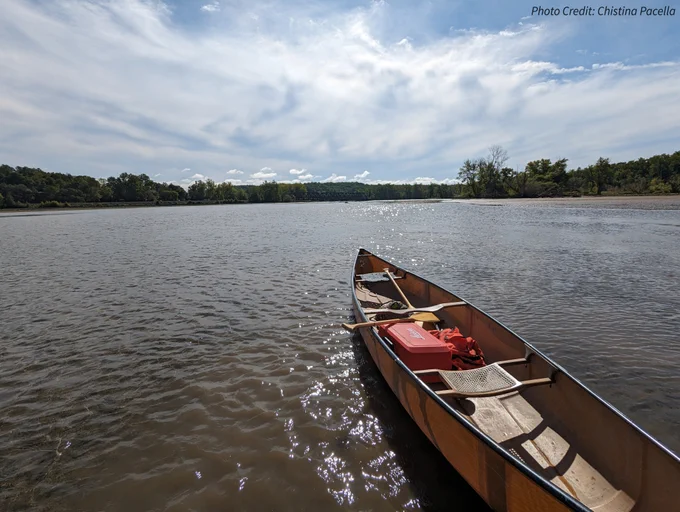 A canoe holding equipment floats on the water pointing towards land and trees in the distance.