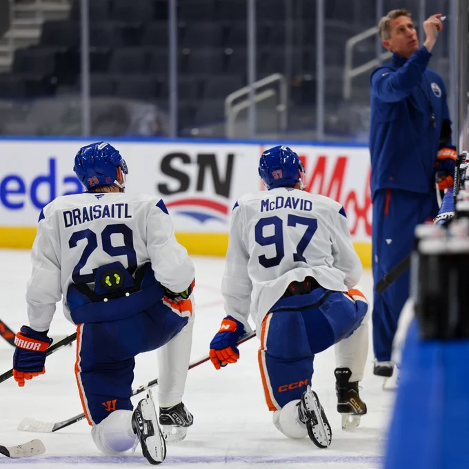 Draisaitl and McDavid kneeling on the ice listening to Coach Knoblauch at Rogers Place. 