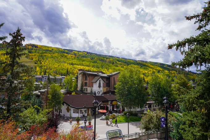 View of Vail, Colorado, featuring a quaint mountain town surrounded by hillsides covered in vibrant yellow aspen trees, under a partly cloudy sky.