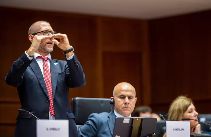 Humberto Insolera, a tall men in a suit, stands up and signs from the podium of the European Parliament