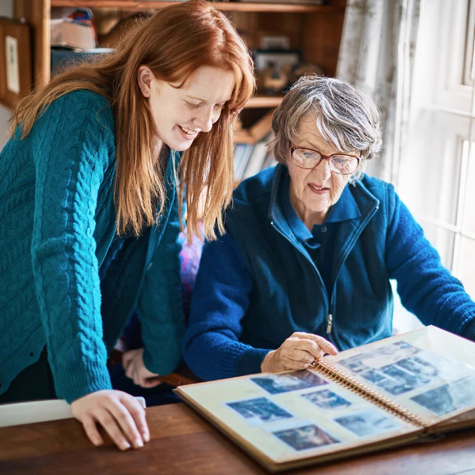 Lady siting at a dining table looking through photo album with adult daughter standing next to her, smiling