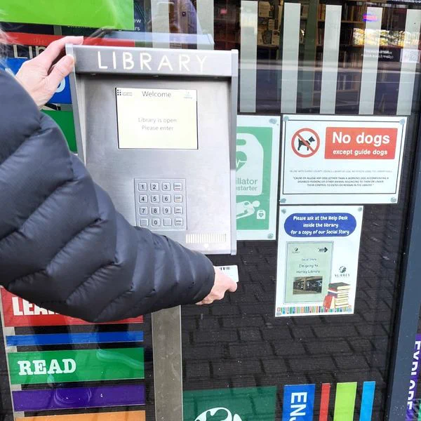 Photo of a person scanning their library card under a machine which says "LIBRARY" across the top. There is a number pad in the middle of the machine. There are some posters in the background