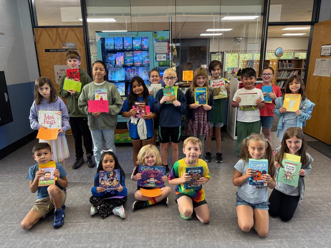17 students gather in front of the book vending machine located outside of the media center. 6 are sitting/kneeling and the rest are standing. they are all proudly holding their new books and certificates. 