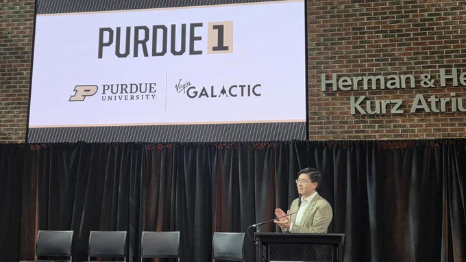 President Mung Chiang speaking at a podium, with a large screen behind them featuring the logos of Purdue University and Virgin Galactic, with Purdue 1 text above.