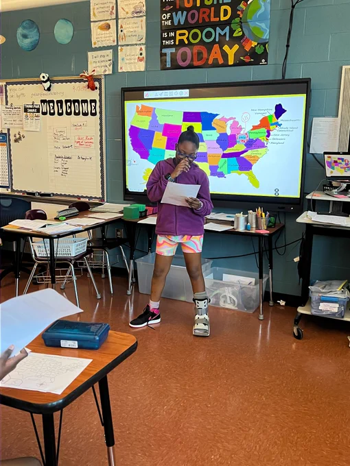 black middle schooler appears shy and laughing as they read their match card of a stem pen pal. purple zip up hoodie and multicolored shorts