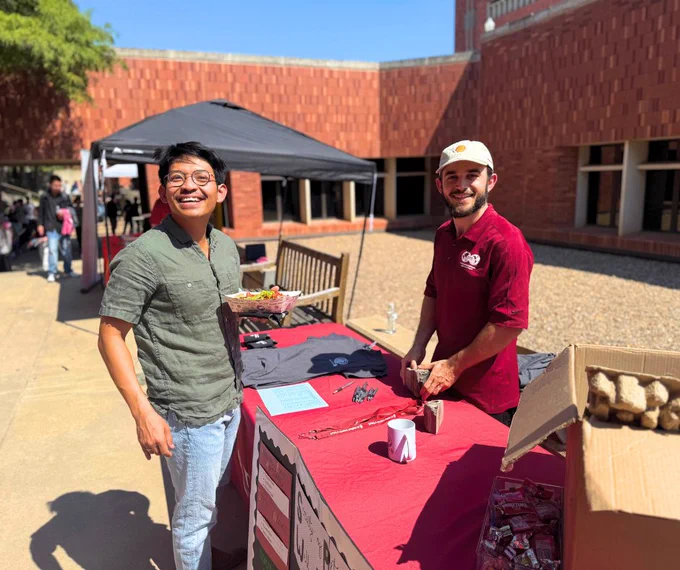 Two individuals staffing a promotional booth at an outdoor event, smiling and standing behind a table covered with informational materials and promotional items, under a banner with the OU logo.