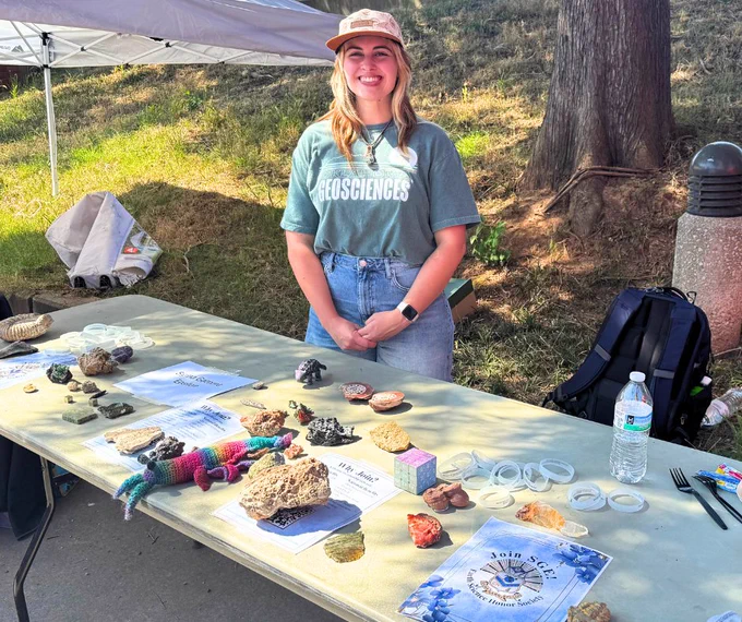 Person standing behind a table displaying various geological samples and educational materials, at an outdoor event with a Geosciences banner.