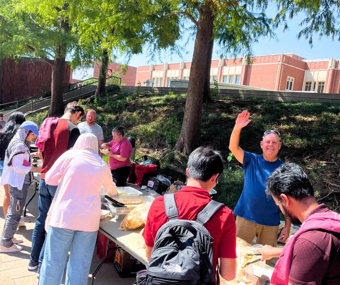 Group of people gathered around a food table outdoors on a sunny day, with one person waving at the camera. Trees and a brick building are visible in the background.