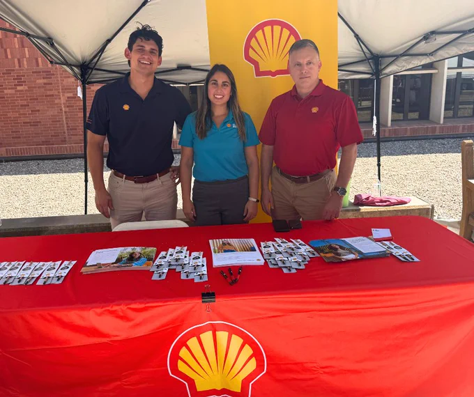 Three individuals standing behind a promotional table with a Shell logo, brochures, and promotional materials displayed on it.