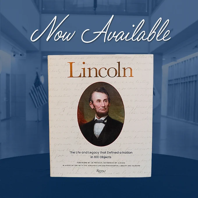 The book Lincoln: The Life and Legacy that Defined a Nation in 100 Objects stands upright on a blue surface. The cream-colored cover features an oval portrait of Abraham Lincoln with handwritten script faintly in the background. Above the book, large text reads “Now Available.” The scene is set inside the Abraham Lincoln Presidential Library and Museum, with flags and architectural details visible in the background.