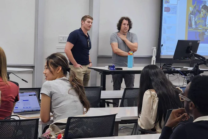 Two instructors standing at the front of a classroom with students working on laptops at AI.Xperience.