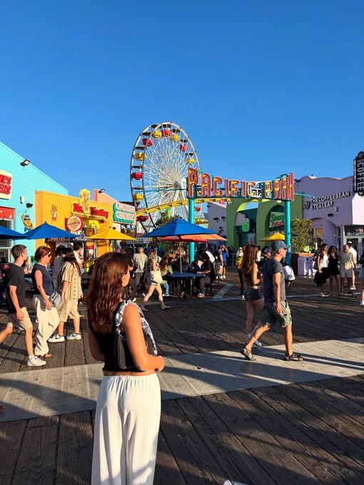 A bustling scene at Pacific Park on the Santa Monica Pier with people walking around, shops, and a large Ferris wheel in the background.