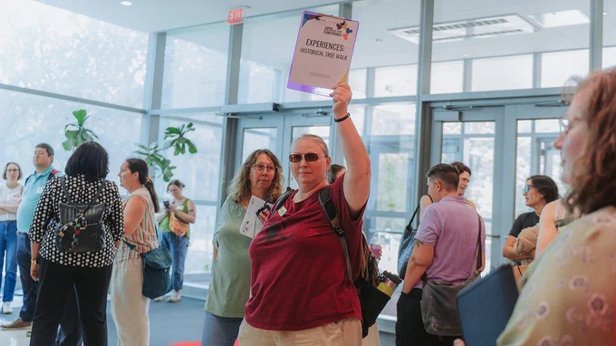A woman holds up a clipboard that reads "Experiences: Historical Tree Walk" as about a dozen other people mill around inside the entrance of a building.