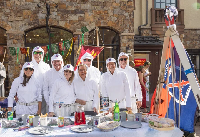 Group of people in 10th Mountain Division costumes smiling behind a food stall decorated with festive banners and flags, at an outdoor event.