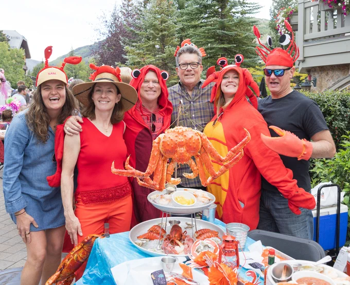 Group of individuals at a festive outdoor event, wearing lobster-themed hats and costumes, posing behind a table with a large crab and themed food.