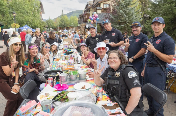 Group of people enjoying a community outdoor gathering around a dining table, with several firefighters and a police officer interacting cheerfully with attendees. There are balloons and a festive atmosphere.