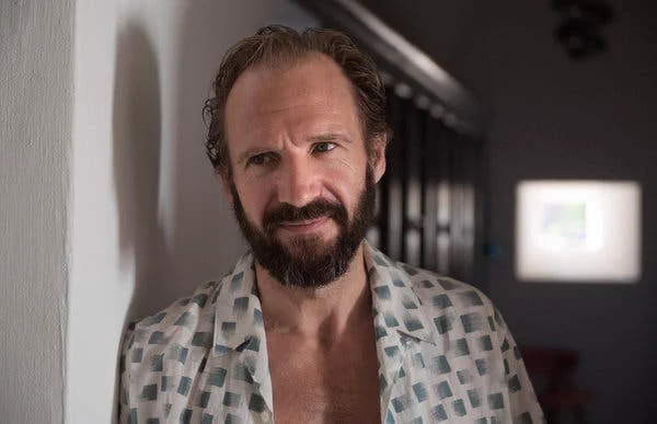 Ralph Fiennes as Harry. He is wearing an unbuttoned white and green patterned shirt, has a thick dark beard, and dark hair with a receding hairline. 