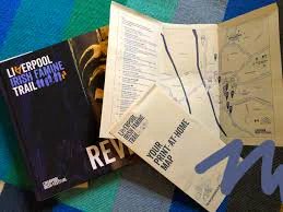 Opened travel guidebook titled "Liverpool Irish Famine Trail" alongside a map and a pamphlet titled "Your Trip Planner," all spread out on a blue textured surface.