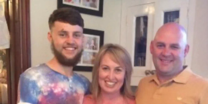 Three people standing closely together in a warmly lit room with framed photos on the wall behind them.