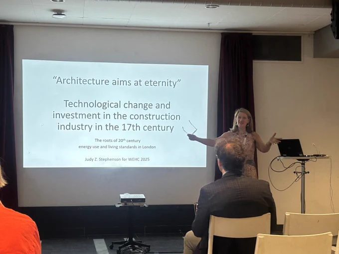 Prof Judy presenting a slideshow titled "Architecture aims at eternity: Technological change and investment in the construction industry in the 17th century,". The audience is visible in the foreground.