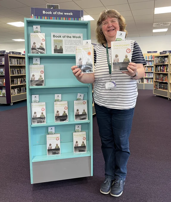 Photo of a sandy haired woman in jeans and a stripy top holding 2 books and standing in front of a pale blue bookcase in a library. The bookcase is displaying 4 rows of the same book. The header at the top of the bookcase reads "Book of the Week" in white lettering on a dark blue background. 