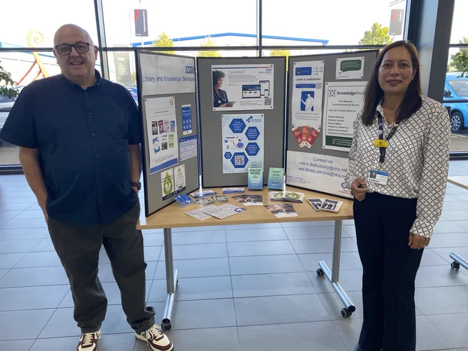 Two individuals, Tommy Whitelaw and Bali Moore, standing at a conference booth at the HCSW / CSW conference. The booth features a tri-fold display with information about the NHS Library and Knowledge Services, including the DynaMed app, KnowledgShare and various promotional materials. The table in front of the display holds leaflets and brochures. The setting is indoors with large windows, showing a car and some outdoor scenery in the background. Tommy is wearing a dark shirt and trousers with white sneakers, while Bali is dressed in a patterned blouse and dark trousers, with a name badge and lanyard.