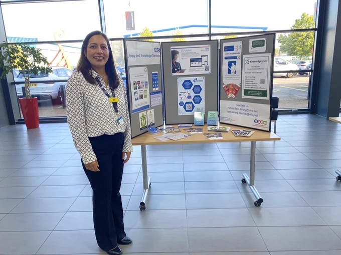 Bali Moore, Senior Library Technician, standing at a conference booth at the HCSW / CSW conference. The booth features a tri-fold display with information about the NHS Library and Knowledge Services, including the DynaMed app, KnowledgShare and various promotional materials. The table in front of the display holds leaflets and brochures. The setting is indoors with large windows, showing a car and some outdoor scenery in the background. Bali is dressed in a patterned blouse and dark trousers, with a name badge and lanyard.
